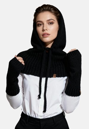 Capa con capucha negra de punto con textura acanalada, mangas largas y cordón. Combinada con una camisa blanca y pantalones negros.