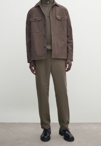 Braune Baumwolljacke mit zwei Brusttaschen, olivfarbener Rollkragenpullover, passende Hose und schwarze Schnürschuhe, vor einem neutralen Hintergrund.