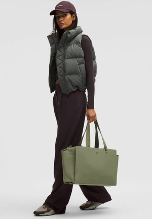 Vesta căptușită de culoare verde închis cu guler înalt, cămașă cu mâneci lungi, pantaloni largi de culoare închisă, geantă tote verde și adidași în tonuri neutre.