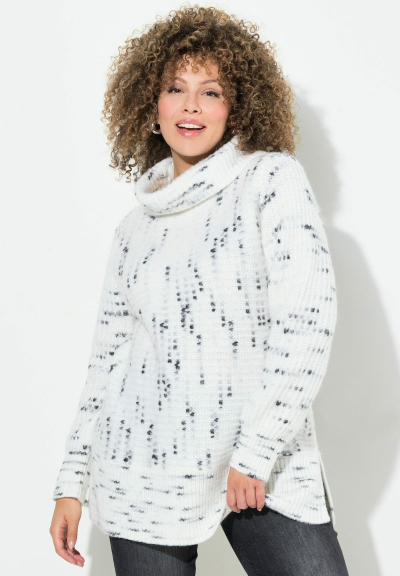 Weißer Pullover mit schwarzem gesprengtem Muster, gestrickter Textur und seitlichen Schlitzen. Kombiniert mit dunklen Jeans.