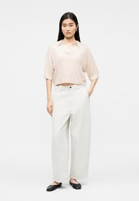 Γυναίκα που φοράει ένα χαλαρό ανοιχτό μπεζ τοπ με μισά μανίκια, φαρδιά άσπρα παντελόνια με ψηλή μέση και μαύρες μπαλαρίνες, όρθια μπροστά σε απλό φόντο.