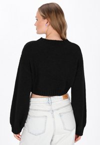 Eine Frau mit hellbraunen Haaren trägt einen schwarzen cropped Pullover mit Ballonärmeln und hochgeschnittenen hellblauen Jeans. Sie steht mit dem Rücken zu einem weißen Hintergrund.
