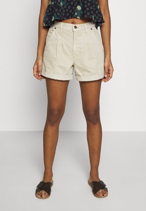 Personne portant un short beige taille haute avec ourlet roulé, un crop top floral noir et des sandales noires, se tenant devant un fond uni.