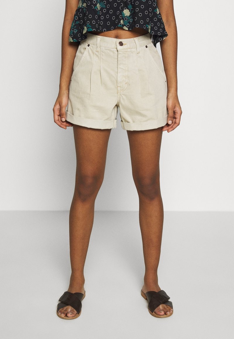 Personne portant un short beige taille haute avec ourlet roulé, un crop top floral noir et des sandales noires, se tenant devant un fond uni.