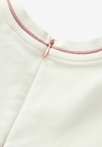 Bílý oděv s hladkou strukturou, který má na zadní straně růžový zip, lemovaný odpovídajícím růžovým látkovým lemem.