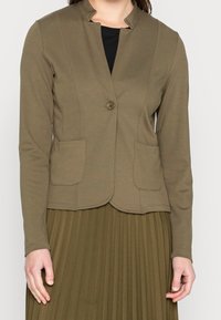 Olive green soft fabric blazer with a rounded collar, single button closure, and two front pockets. Worn over a pleated skirt.