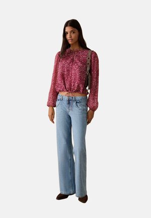 Blusa roja con patrón de cachemira y cintura con cordón, combinada con vaqueros azul claro de pierna ancha. La modelo lleva zapatos marrones y detalles mínimos en accesorios.