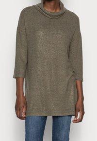 Olivgrünes Strickoberteil mit einem Schalkragen, Dreiviertelärmeln und lässiger Passform, kombiniert mit hellblauen Jeans.