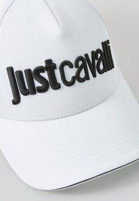 Hvit baseballcaps med brodert svart "Just Cavalli" tekst, med en strukturert form og flat skygge. Teksturert stoff med syddede detaljer.