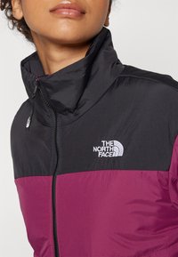 Sort og lilla jakke med høj krave, halv lynlås lukning, og "The North Face" logo i hvidt på brystet. Letvægts, tekstureret stof.