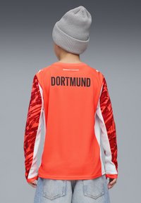 Kirkas oranssi urheilu-paita, jossa on punaiset kuvioidut hihat, valkoiset yksityiskohdat ja "DORTMUND" painettuna selässä paksuilla mustilla kirjaimilla.