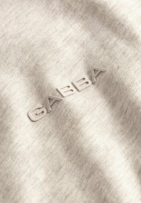 Beiges geripptes Gewebe mit erhabenen, hellfarbenen "GABBA"-Markenbuchstaben, die auf dem Material geprägt und zentriert sind.