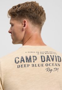 Light beige t-shirt with a round neckline and short sleeves. The back features dark blue text reading "CAMP DAVID DEEP BLUE OCEAN."