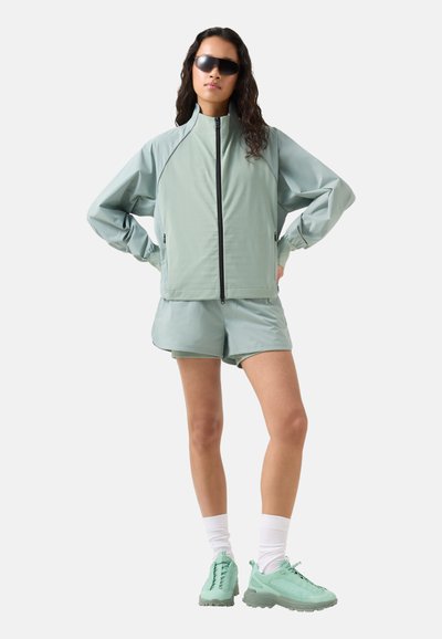 Kvinde iført mintgrøn vindjakke, matchende shorts, hvide sokker, mintgrønne sneakers og sorte solbriller, stående med hænderne på hofterne.