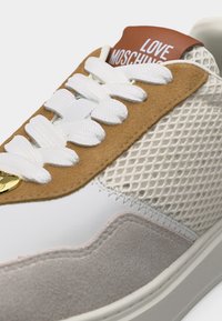 Λευκό αθλητικό παπούτσι με μπεζ σουέντ, πλαϊνά από κρεμ μέσχ, λευκά κορδόνια, καφέ γλώσσα με την ετικέτα "LOVE MOSCHINO" και χρυσό κρεμαστό καρδιάς στην άκρη του κορδονιού.