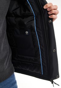 Giacca nera imbottita con piping interno blu e tasca per etichetta. Presenta una chiusura con zip e bottoni a pressione. Tessuto morbido e design elegante.