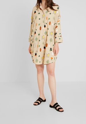 Kvinde iført en beige skjortekjole med farverige abstrakte blomster- og geometriske mønstre, kombineret med sorte åbne sandaler.