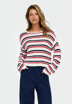 Vrouw draagt een witte gebreide trui met rode en zwarte horizontale strepen en marineblauwe broek met hoge taille, staat met handen in de zakken.