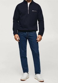 Pullover blu navy con collo a zip in tessuto morbido con tasca frontale, abbinato a jeans blu e sneaker bianche. Design semplice, casual e vestibilità comoda.