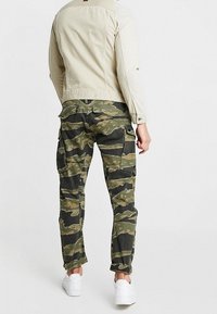 Mężczyzna w beżowej kurtce, zielonych i czarnych spodniach cargo w kamuflażu oraz białych trampkach, stojący tyłem do przodu.