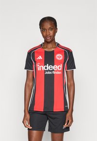 Juoda ir raudona dryžuota futbolo marškinė su apvaliu kaklu, baltais akcentais ir „Indeed Jobs finden“ logotipu. Dėvima su juodomis šortais.