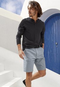 Maglietta henley nera a maniche lunghe con bottoni, pantaloni corti azzurri, cintura nera e scarpe nere, posato davanti a una parete bianca e una porta blu.