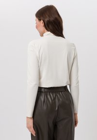 Long-sleeve, cream top with a high neck, made of smooth fabric. Paired with high-waisted, dark brown, pleated leather pants.