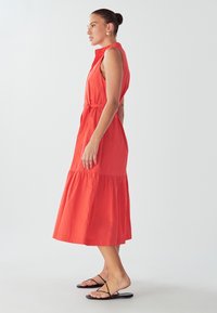 Rdeča midi obleka brez rokavov, izdelana iz gladkega blaga s plastehnim krilom, gumbi in pasom za zavezovanje v pasu. Oblečena z črnimi sandali.