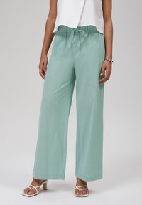 Pantalones de pierna ancha en verde menta claro, hechos de una tela texturizada, con cinturilla elástica y cordón, combinados con sandalias blancas de tiras.