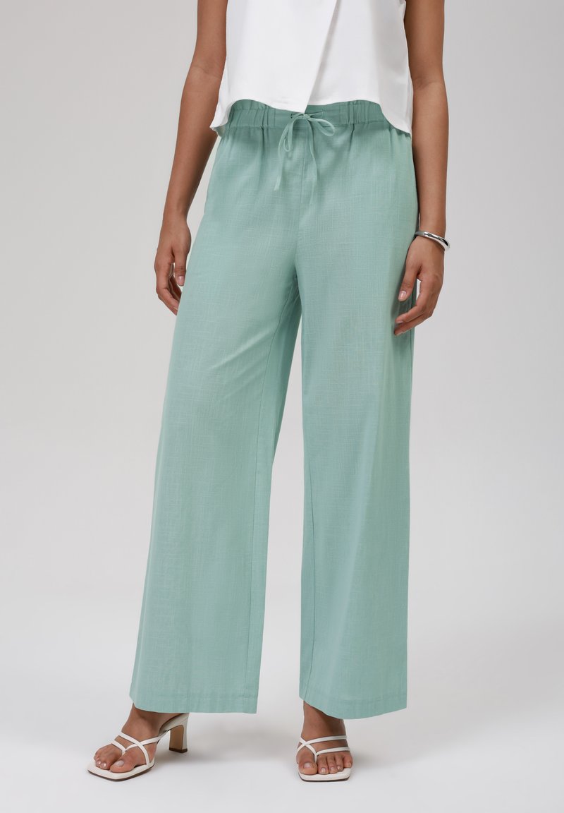Pantalones de pierna ancha en verde menta claro, hechos de una tela texturizada, con cinturilla elástica y cordón, combinados con sandalias blancas de tiras.