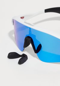 Occhiali da sole sportivi con montatura bianca, lenti a gradiente blu e cuscinetti nasali in gomma nera. Include anche ulteriori supporti per le orecchie in gomma nera.