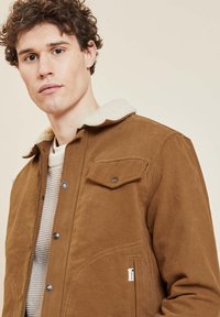 Chaqueta de pana marrón con un suave cuello de piel sintética color crema, que cuenta con botones a presión y un bolsillo en el pecho del lado izquierdo.