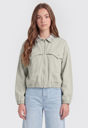 Junge Frau mit langen, welligen braunen Haaren, die eine hellbeige Reißverschlussjacke mit karierter Kragen und hellblaue Jeans trägt, steht vor einem einfarbigen Hintergrund.