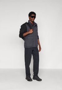 Schwarze Steppweste mit Reißverschlusstaschen, getragen über einem langärmligen schwarzen Hemd. Schwarze Hose und Schuhe ergänzen das Outfit. Dunkle Sonnenbrille inklusive.