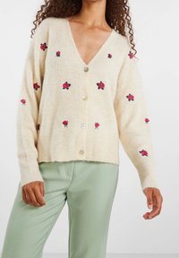 Krämfärgad stickad cardigan med V-ringning, knäppning framtill och broderade rosa rosor. Kombinerad med ljusgröna byxor. Mjuk textur.