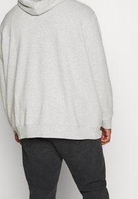 Una persona che indossa una felpa grigia chiara e jeans grigi scuri, vista da dietro su uno sfondo bianco semplice.