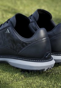 Chaussures de sport noires avec semelles blanches reposant sur de l'herbe verte, montrant les détails arrière et latéraux avec de petits logos triangulaires.
