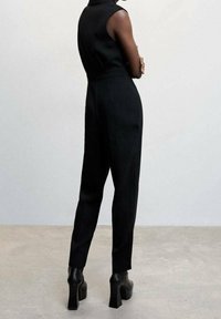 Black sleeveless jumpsuit with tapered legs, smooth texture, high neckline, and visible seams, paired with chunky black heeled boots.