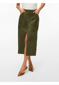 Femme portant une jupe midi vert olive à fente frontale et poches, associée à des escarpins slingback pointus marron, posant devant un fond blanc.