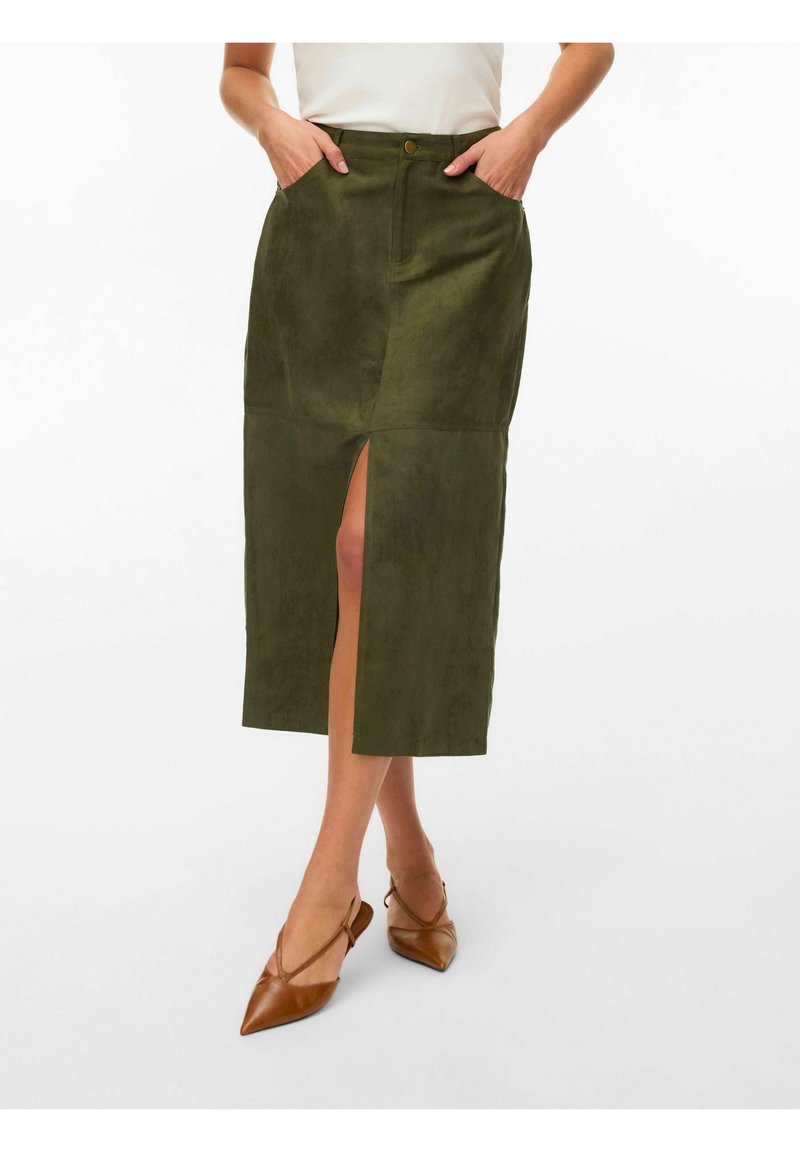 Femme portant une jupe midi vert olive à fente frontale et poches, associée à des escarpins slingback pointus marron, posant devant un fond blanc.