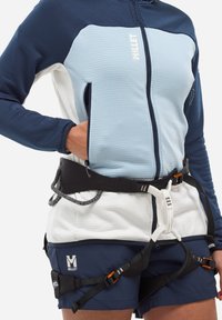 Person wearing a blue and white zip jacket and blue shorts, fitted with a black and white climbing harness with orange buckles.