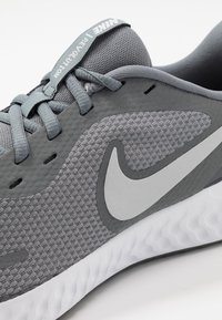 Šedá sportovní obuv s texturovaným síťovým svrškem, šedými tkaničkami, bílým logem ve tvaru klíče a bílou polstrovanou podrážkou se vlnitým vzorem.