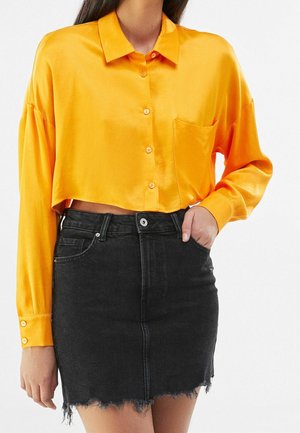 Camisa de satén naranja corta con cuello, cierre de botones en la parte delantera y un bolsillo, combinada con una falda de mezclilla negra con dobladillo deshilachado.