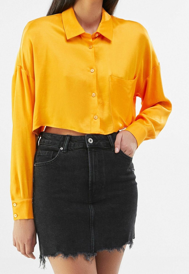 Satin, kort orange skjorta med krage, knappframsida och en ficka, parat med en svart denimkjol med fransad fåll.