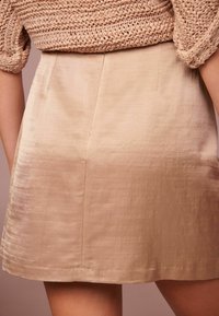 Une jupe beige, longueur genoux, en tissu lisse avec un léger éclat. Elle présente une coupe ajustée et des détails de couture simples.