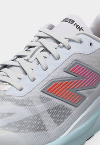 Λευκό αθλητικό παπούτσι με λευκά κορδόνια, γκρι και πορτοκαλί λογότυπο "N" στο πλάι και ανοιχτό μπλε σόλα, κοντινή άποψη του εμπρόσθιου πάνω μέρους.