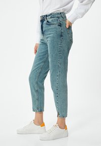 Ljusblå denimjeans med hög midja, tapered passform, frampockets och dragkedjor vid anklarna. Matchas med vita sneakers med orangea accenter.