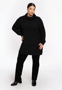 Übergroßer schwarzer Pullover mit Kapuze und strukturiertem Strickmuster, kombiniert mit schwarzen, eng anliegenden Hosen und schwarzen Stiefeletten mit Absatz.
