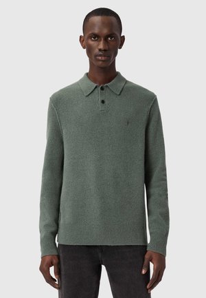 Mies, jolla on vihreä pitkähihainen poolopaita mustilla napeilla ja tummat housut, seisoo vaaleanharmaan taustan edessä.