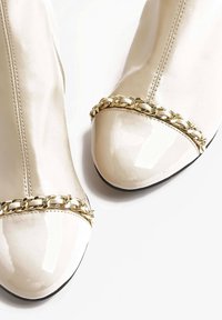 Lakované kotníkové boty v krémové barvě, s lesklou špičkou a dekorativním zlatým řetízkem kolem kotníku. Celkově hladký povrch.